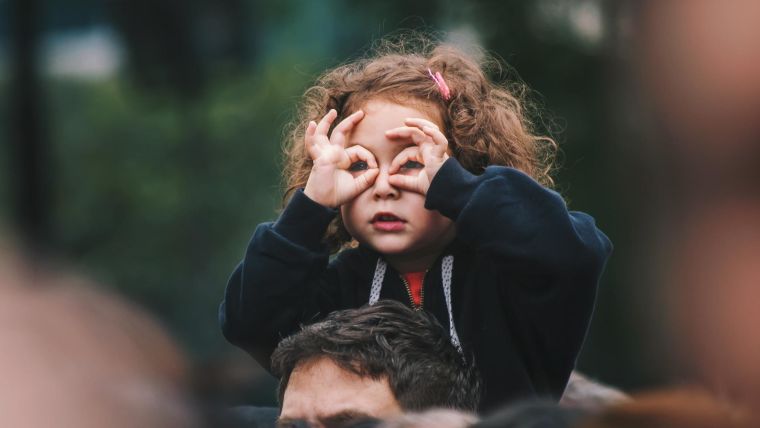 A small girl with curly hair is sitting on a man's shoulders. She is using her hands to form binoculars to look through.