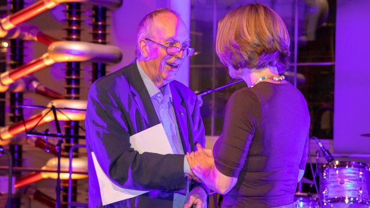 Caroline Struthers shakes hands with Iain Chalmers on a stage. In the background are music stands and a drum kit.